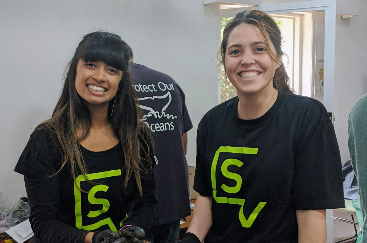 Two young women stand side by side, smiling at the camera. The girl on the left has dark hair and a fringe. The girl on the right has brown hair in a ponytail. Both wear black t-shirts with a light green logo in the centre.
