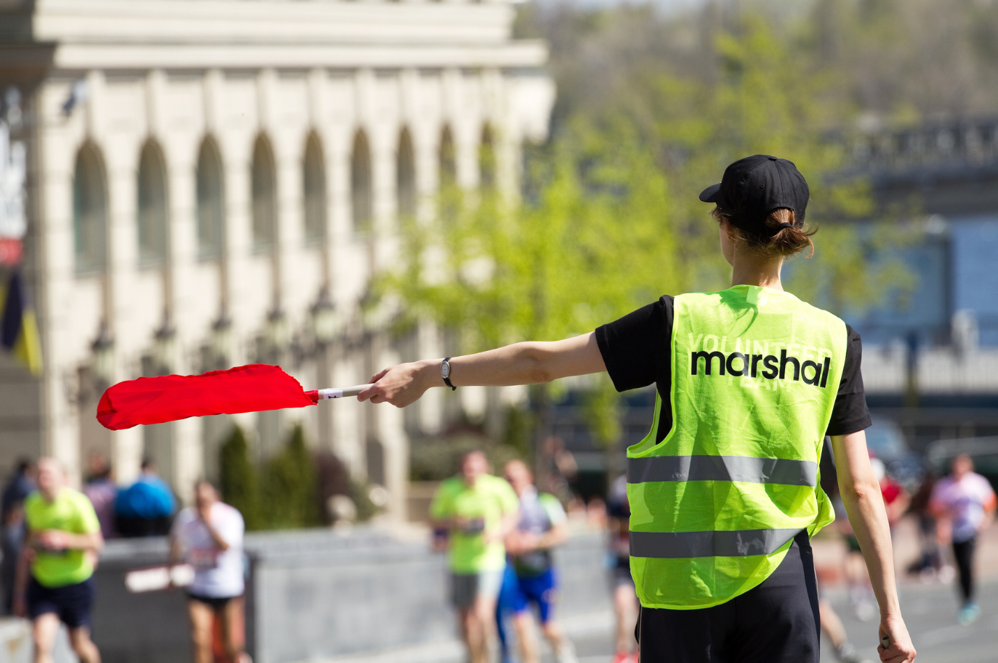 A volunteer wearing a black t-shirt, black cap and a yellow high-vis vest with 'volunteer marshal' written on the back. Her left arm is out to the side, holding a red flag to direct runners on a course.