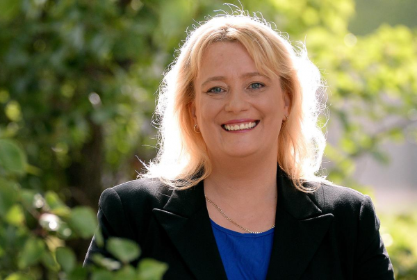 Ros Spence is standing in front of some outdoor greenery. She has shoulder-length blonde hair and is wearing a blue top underneath a black blazer.