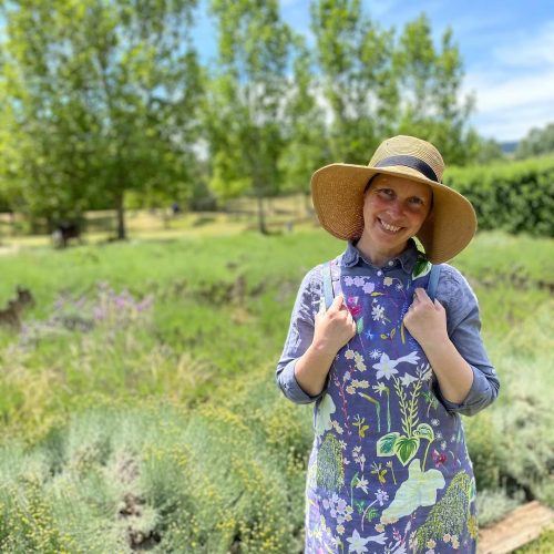 Paula standing in a green garden, wearing a broad hat and purple dress with flowers