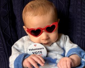 A baby girl wearing red heart-shaped sunglasses and a badge that says 'Volunteers Vote'.