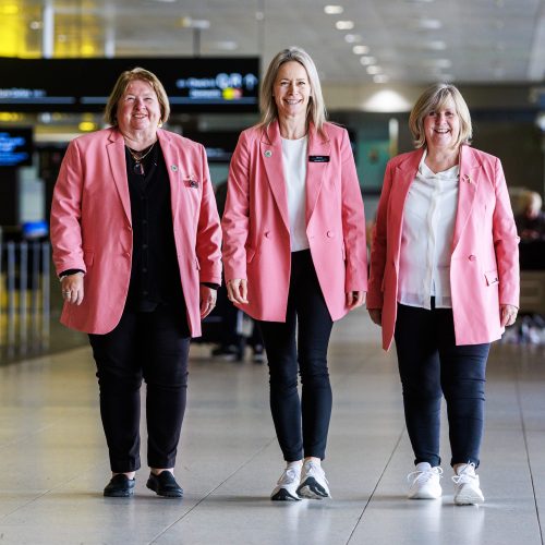 Three Melbourne Airport Experience Ambassadors in bright melon blazers, walking the terminal at Melbourne Airport